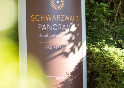 A billboard with text on it pointing towards the entrance of the hotel SCHWARZWALD PANORAMA. Text: "SCHWARZWALD PANORAMA – Hotel. Campus. Selfness. Bad Herrenalb – 100m -->"