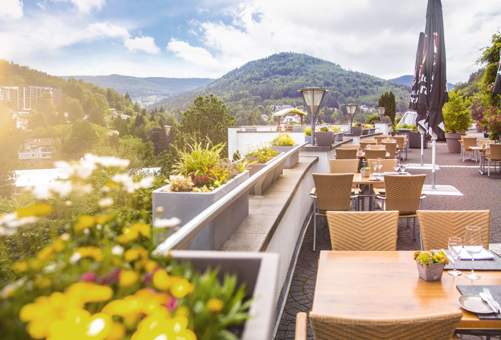 Auf einer Dachterasse stehen Tische und Stühle, im Hintergrund sieht man Wälder und die Andeutung einer Stadt, die Aussicht der Hotel Schwarzwald Panorama